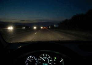 Driver’s point of view on a dark highway with blurred lane markings, dim road signs, and headlight glare caused by cataracts and night driving difficulty.
